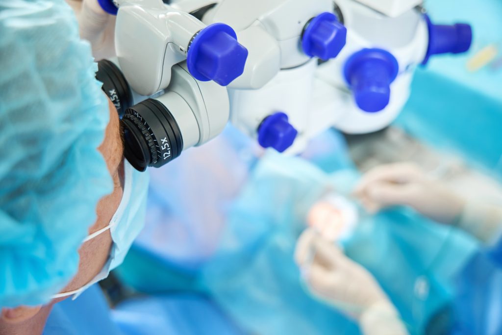 Doctor in protective , mask and hat looking in surgical microscope and looking through it on patient